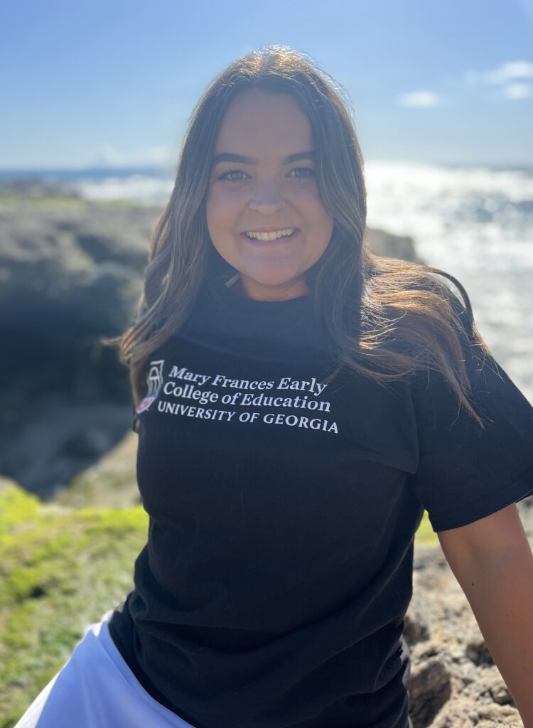 Courtney Adams sits on a cliff overlooking the ocean, wearing a UGA Mary Frances Early College of Education shirt